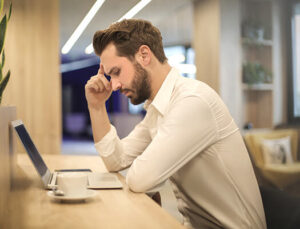 man in tan shirt with cup of coffee sitting at desk looking at computer